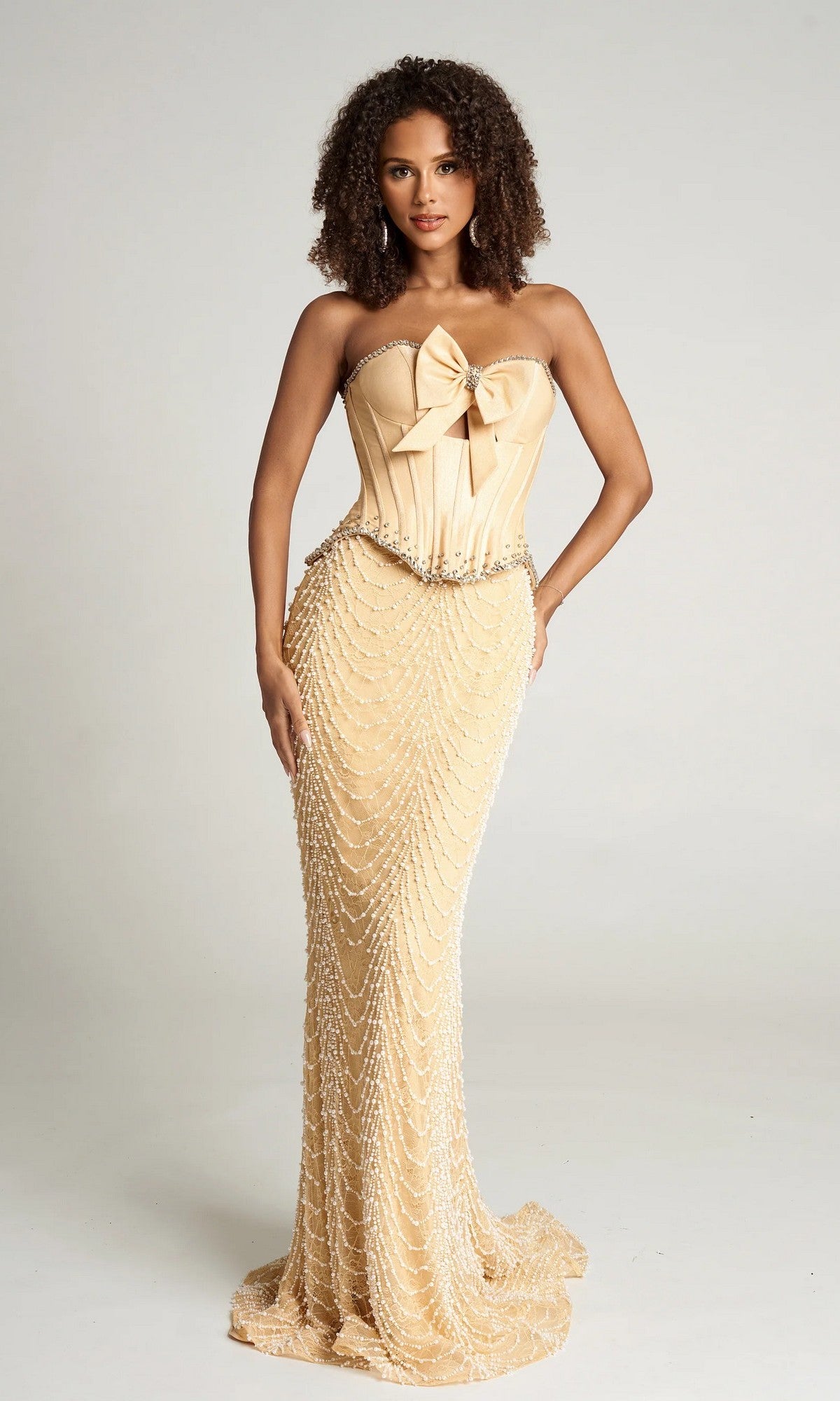 A woman wears the Portia and Scarlett PS26054 long formal dress in beige, featuring a strapless corset bodice, a large bow accent, and beaded draping details, posing against a plain background.